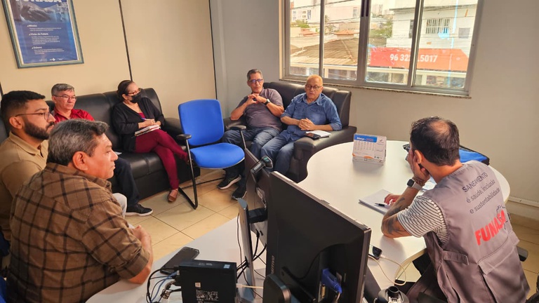 Close do grupo em reunião, com foco em uma pessoa apontando para documentos sobre a mesa enquanto propõe o uso de um laboratório móvel de análise de água.