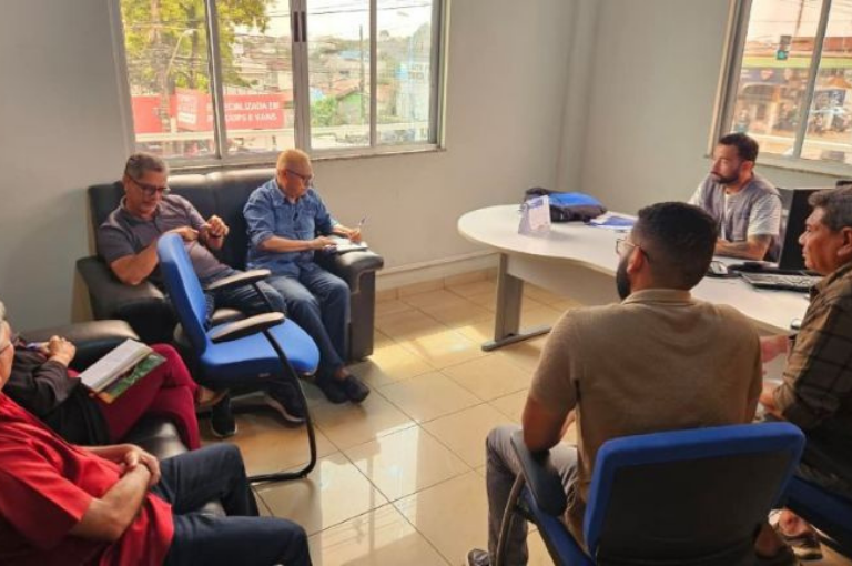 Seis profissionais aparecem reunidos em torno de uma mesa com notebooks e documentos, em uma sala de reuniões, discutindo cooperação técnica.