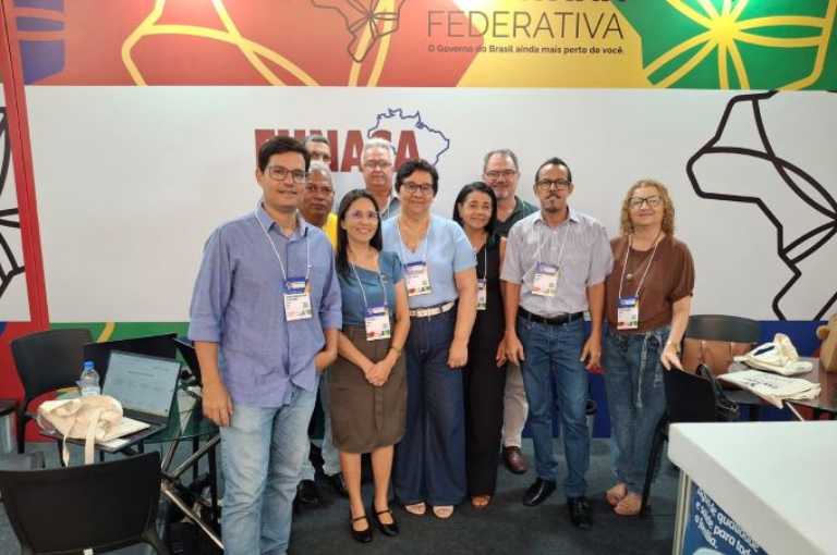 Foto em grupo com dez pessoas (homens e mulheres) posando sorridentes em frente a um painel colorido da "Caravana Federativa" com o logotipo da Funasa.