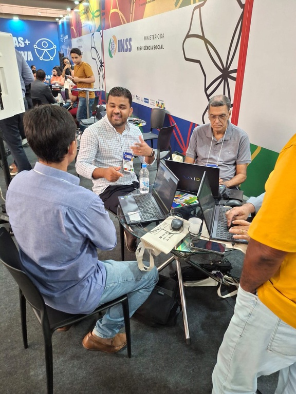 Três homens aparecem sentados ao redor de uma mesa de vidro com vários notebooks, em uma conversa técnica durante o atendimento a gestores.