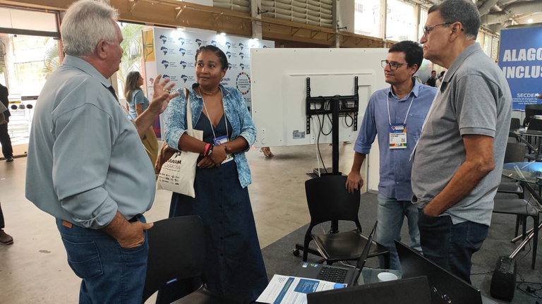 Um homem de camisa clara gesticula enquanto conversa com uma mulher e outros dois homens em pé, em um amplo pavilhão de eventos.