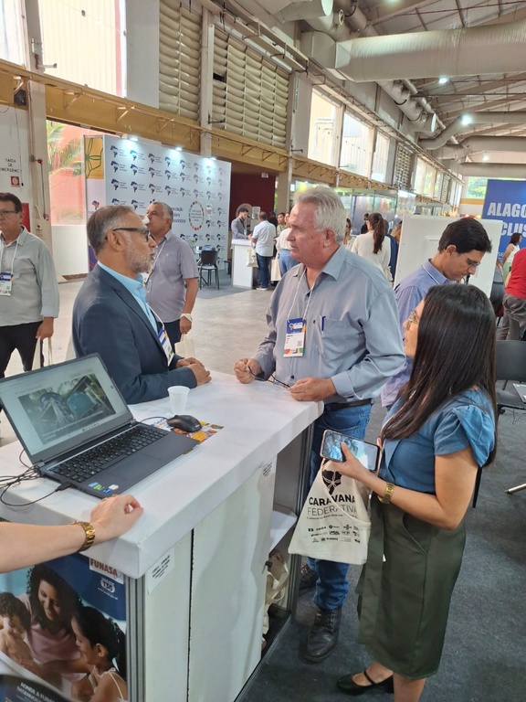 Um homem de terno presta orientações a um casal em um balcão branco, onde um notebook exibe imagens de reservatórios de água.