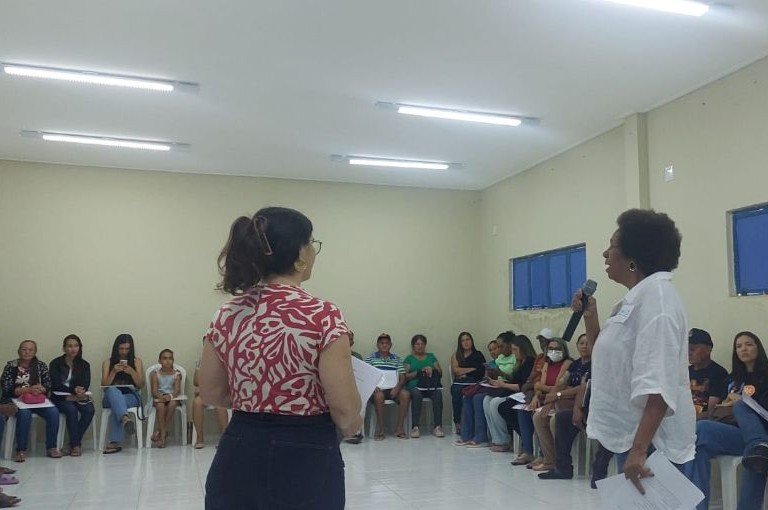 Em uma sala clara, uma mulher fala ao microfone para um público de cerca de 20 pessoas sentadas em cadeiras ao longo das parede.,