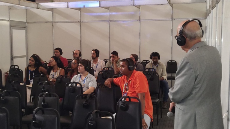 Palestrante de terno cinza e fones fala ao microfone para plateia sentada e atenta, também usando fones, em sala de conferência.