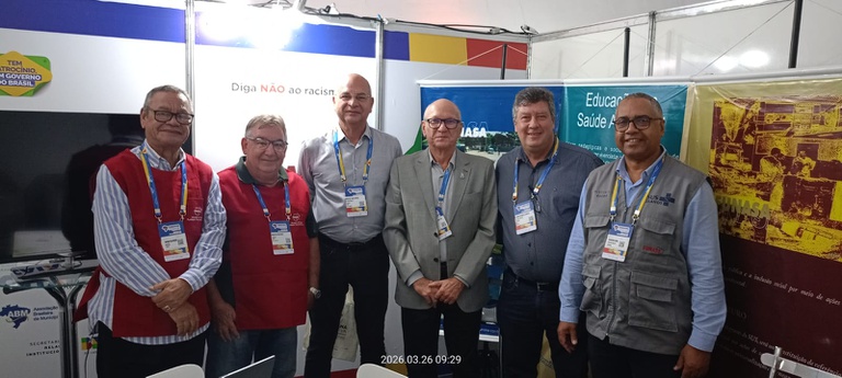 Seis homens com crachás posam para foto em um estande. Dois usam colete vermelho, um colete cinza e três camisa social. Banners ao fundo.