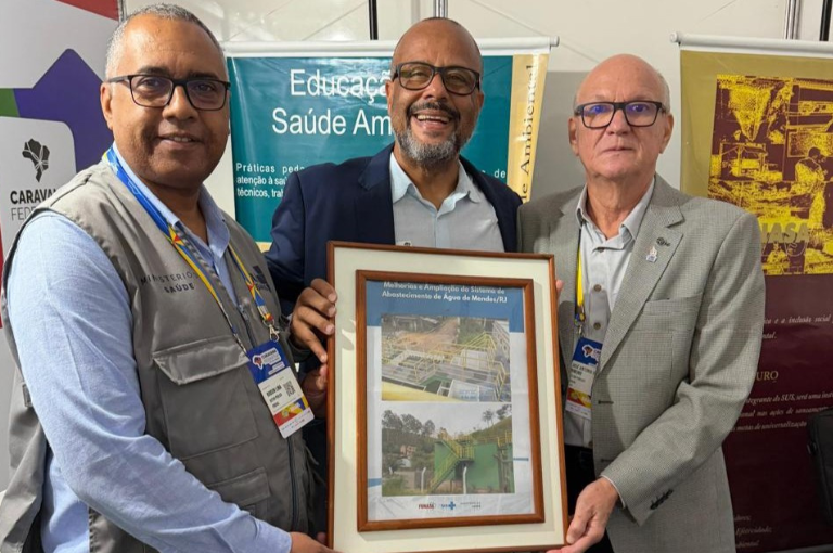 Três homens sorrindo seguram um quadro emoldurado que exibe duas fotos de uma obra de saneamento. Eles estão em um estande de evento.