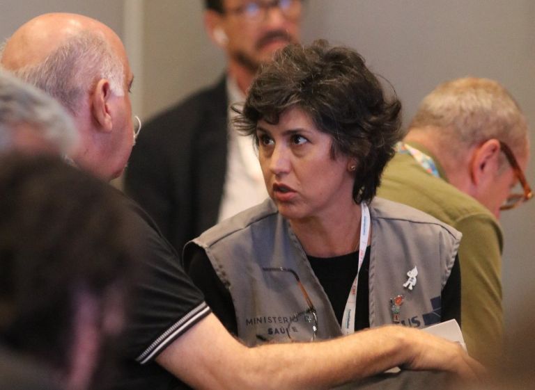 Um close da técnica Luciana Fonseca, vestindo colete do Ministério da Saúde, em conversa atenta com um participante.