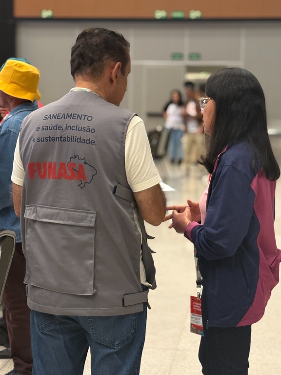 Vista das costas de um servidor da Funasa com o slogan "Saneamento é saúde, inclusão e sustentabilidade" durante diálogo com uma participante.