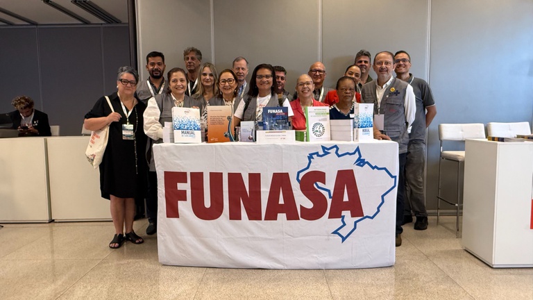 Cerca de 15 profissionais da instituição posam atrás de um balcão branco com o logotipo da Funasa e diversos manuais técnicos sobre saneamento.