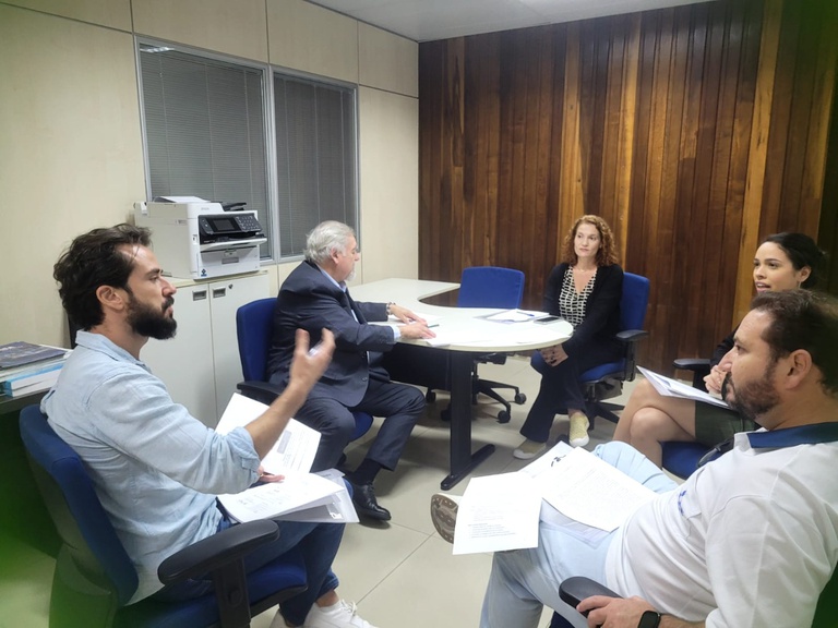 Cinco pessoas sentadas ao redor de uma mesa redonda em escritório, discutindo documentos impressos em reunião técnica.