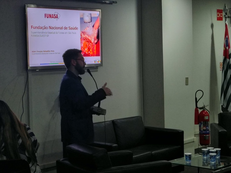 Homem gesticula em frente a monitor com slide da Funasa. Ao lado, poltronas pretas, mesa de centro com copos d'água e extintor.