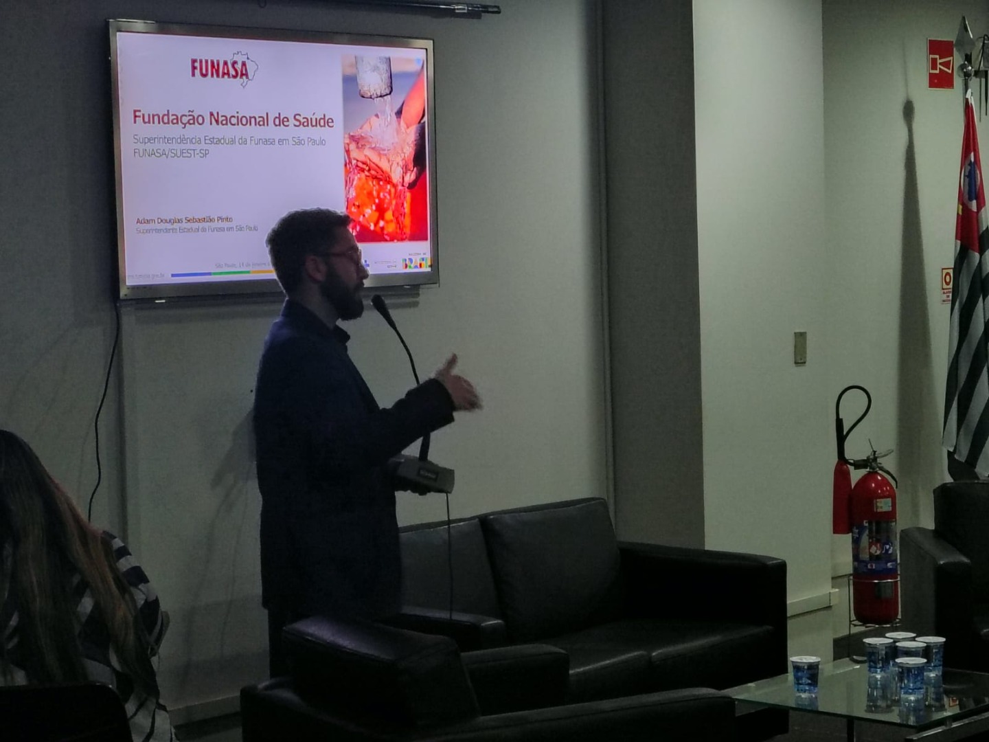 Homem gesticula em frente a monitor com slide da Funasa. Ao lado, poltronas pretas, mesa de centro com copos d'água e extintor.