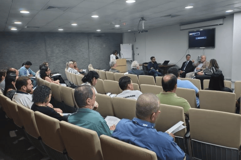 Plateia de costas assistindo a palestra em auditório com painel de especialistas ao fundo e tela de projeção ligada.