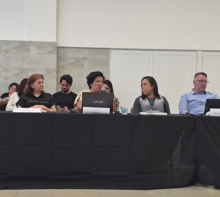 Mesa redonda com cinco integrantes, onde uma mulher ao centro fala ao microfone diante de notebooks em uma mesa com toalha preta.