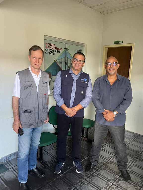 Ademar Zanini, Eduardo Ribeiro e José Renato sorriem em pé em um corredor. Ao fundo, cartaz com a frase: Nossa bandeira é cuidar de gente!