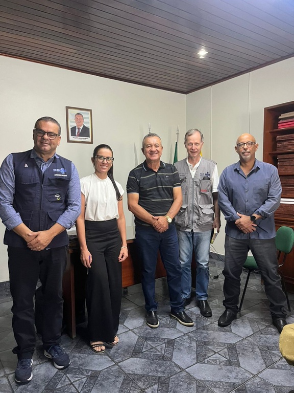 Cinco pessoas em pé em gabinete com teto de madeira. À esquerda, homem com colete azul do SUS; ao lado, mulher de blusa branca e calça preta.