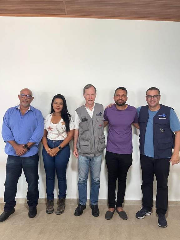 Cinco pessoas em pé, lado a lado, em frente a uma parede branca. Dois homens vestem coletes cinza e azul com logotipos do SUS e do Ministério da Saúde.