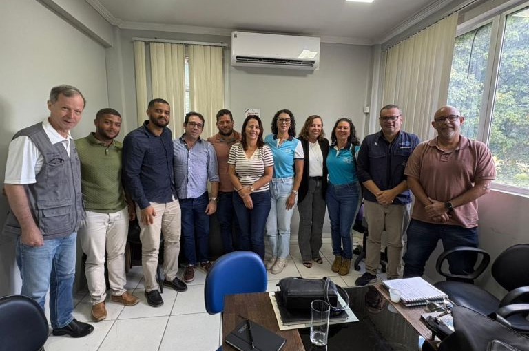 Onze pessoas sorriem em pé em um escritório. À esquerda, homem com colete cinza do Ministério da Saúde. Ao centro, mesa com caderno e copo.