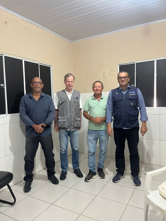Quatro homens em pé em sala de azulejos brancos. Dois vestem coletes oficiais sobre camisas sociais. O segundo à esquerda usa colete cinza.