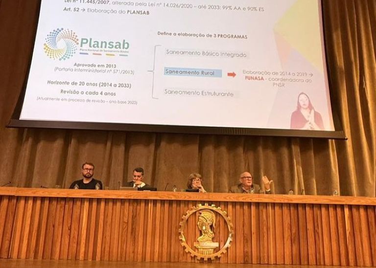 Quatro homens sentados à mesa com microfones; o primeiro da direita fala durante painel técnico sobre saneamento e saúde.
