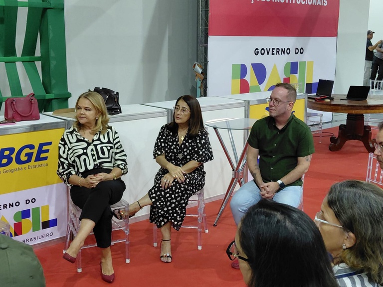 Duas mulheres e um homem sentados em cadeiras transparentes conversam em estande de eventos com tapete vermelho.