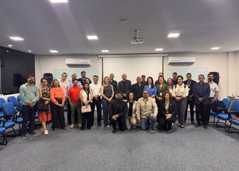 Grupo de cerca de trinta pessoas posa para foto oficial em auditório com projetor e cadeiras azuis ao fundo.