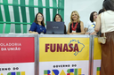 Três mulheres sorriem atrás de balcão da Funasa com notebook. Painel exibe logotipos da Funasa e do Governo do Brasil.