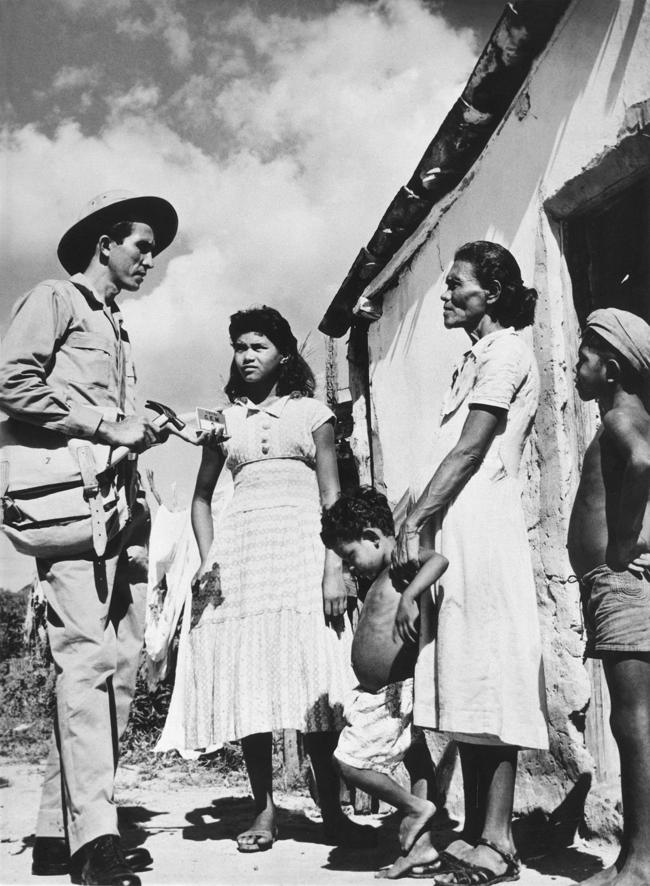 Agente de saúde de chapéu conversa com família em frente a uma casa simples em foto PB.