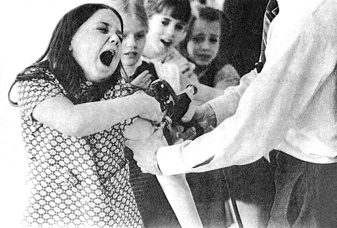 Menina chorando ao receber vacina com injetor de pressão no braço, cercada por outras crianças.
