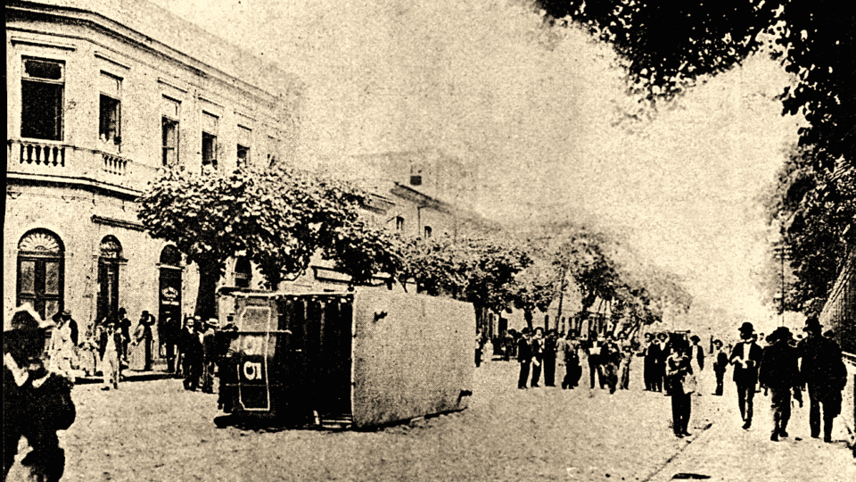 Foto antiga de um bonde virado em rua de paralelepípedos com multidão ao redor celebrando.