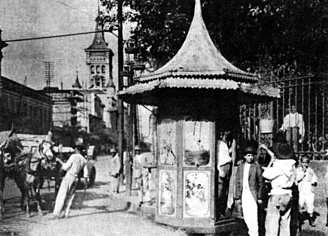 Foto antiga em sépia de homens reunidos ao redor de um quiosque de rua decorado com teto pontiagudo.