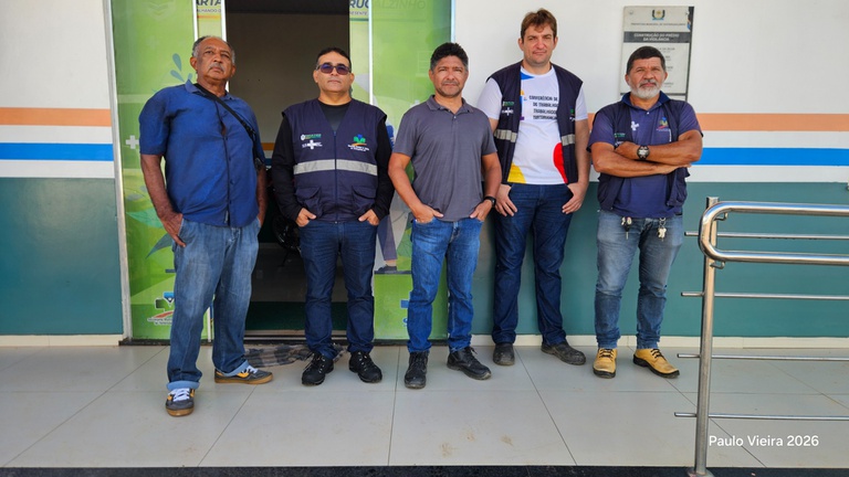 Cinco homens da equipe técnica posam alinhados em frente à entrada de um prédio oficial com faixas coloridas na parede.