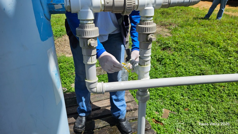 Close de mãos com luvas colhendo amostra de água em um pequeno saco plástico a partir de uma tubulação branca de PVC.