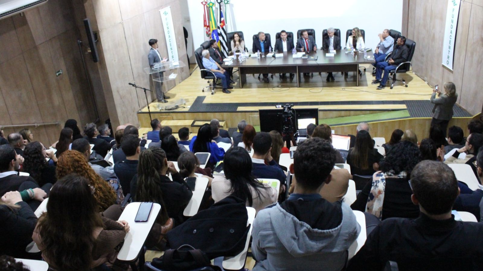 Pra todos verem: foto do auditório lotado com mesa ao fundo.