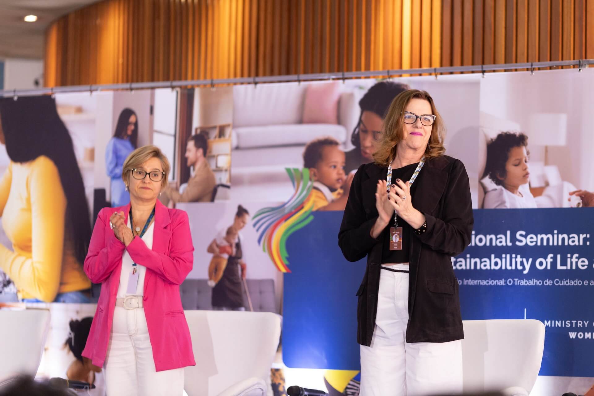 Na imagem, vemos duas mulheres de pé em um palco. Ambas estão aplaudindo e olhando para algo ou alguém fora do quadro. A mulher à esquerda, a secretária nacional Rosane Silva, usa um blazer rosa sobre uma blusa branca e calça branca. Ela tem cabelo curto e usa óculos. A mulher à direita, a secretária-executiva Maria Guarezi, usa um blazer preto sobre uma blusa preta e calça branca. Ela tem cabelo um pouco mais longo e também usa óculos. Atrás delas, há um banner com o título "International Seminar: Sustainability of Life" (Seminário Internacional: Sustentabilidade da Vida) e várias imagens de pessoas em contextos de trabalho e cuidado, incluindo uma mãe com um bebê e profissionais em um escritório.