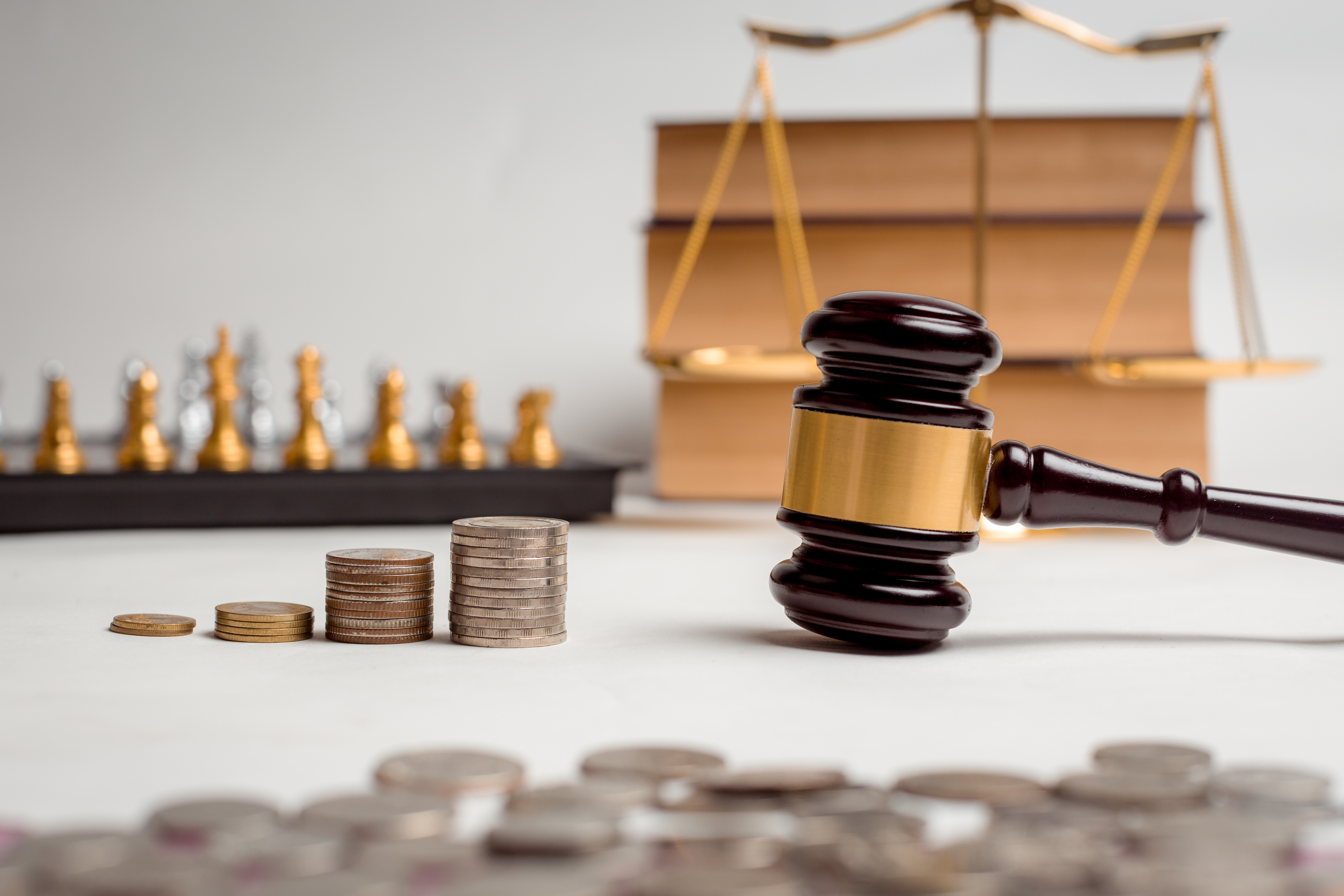 A imagem mostra um martelo de juiz de madeira com uma faixa dourada, colocado sobre uma superfície branca, ao lado de pilhas de moedas em diferentes alturas, sugerindo valores variados. Ao fundo, desfocados, estão uma balança de justiça e livros empilhados, além de peças de xadrez douradas e prateadas alinhadas. A composição parece evocar temas de justiça, economia, decisões judiciais e equilíbrio, simbolizando possivelmente a relação entre finanças e o sistema jurídico.