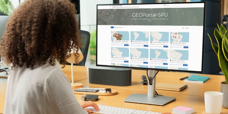 Imagem de uma mulher de cabelo crespo em frente a um computador. Na tela, imagem da tela inicial do Geoportal da SPU.