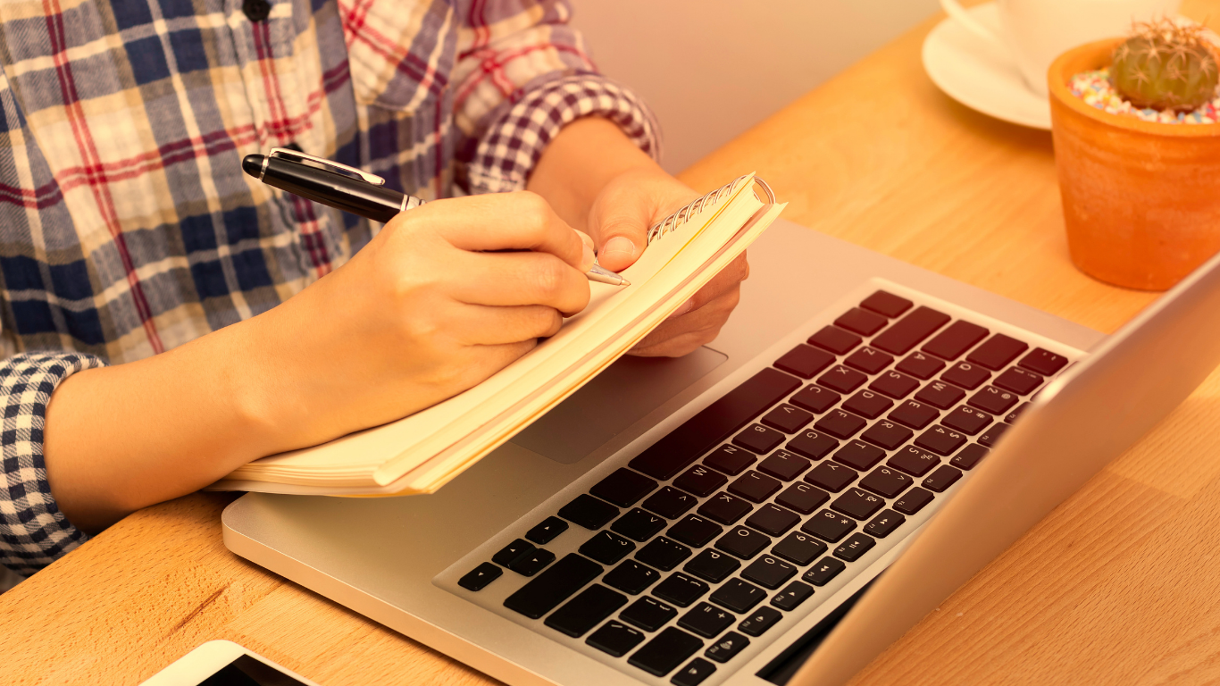 Pessoa escrevendo num caderno em frente a um computador
