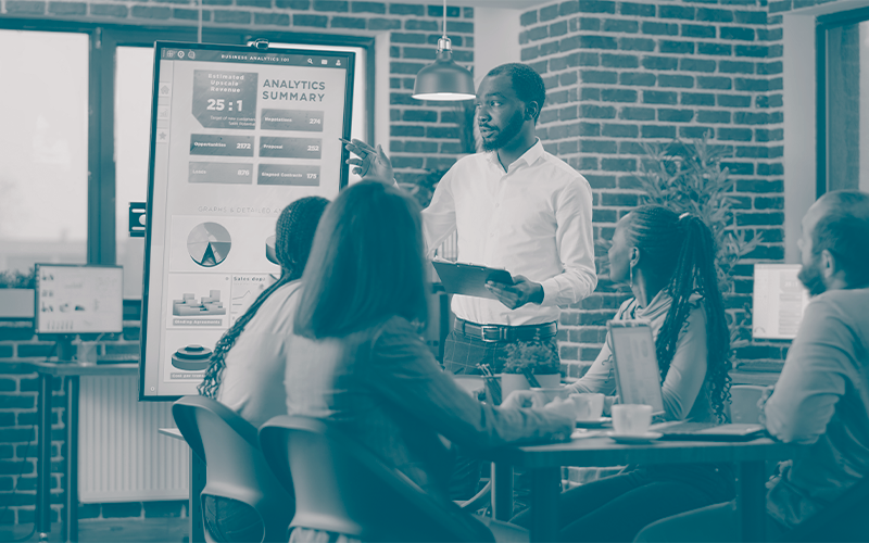 Professor apontando para uma apresentação de dados e quatro pessoas sentadas em volta de uma mesa observando.