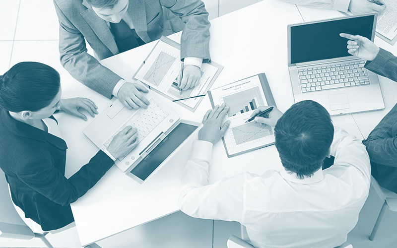 Quatro pessoas reunidas em volta de uma mesa analisando gráficos no notebook e em impressões.