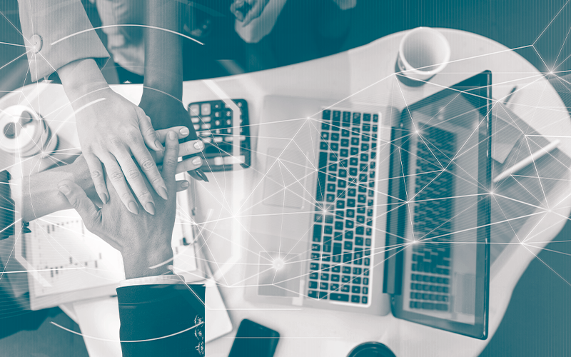 Pessoas colocando uma mão sobre a outra em uma mesa, próximo a um notebook.