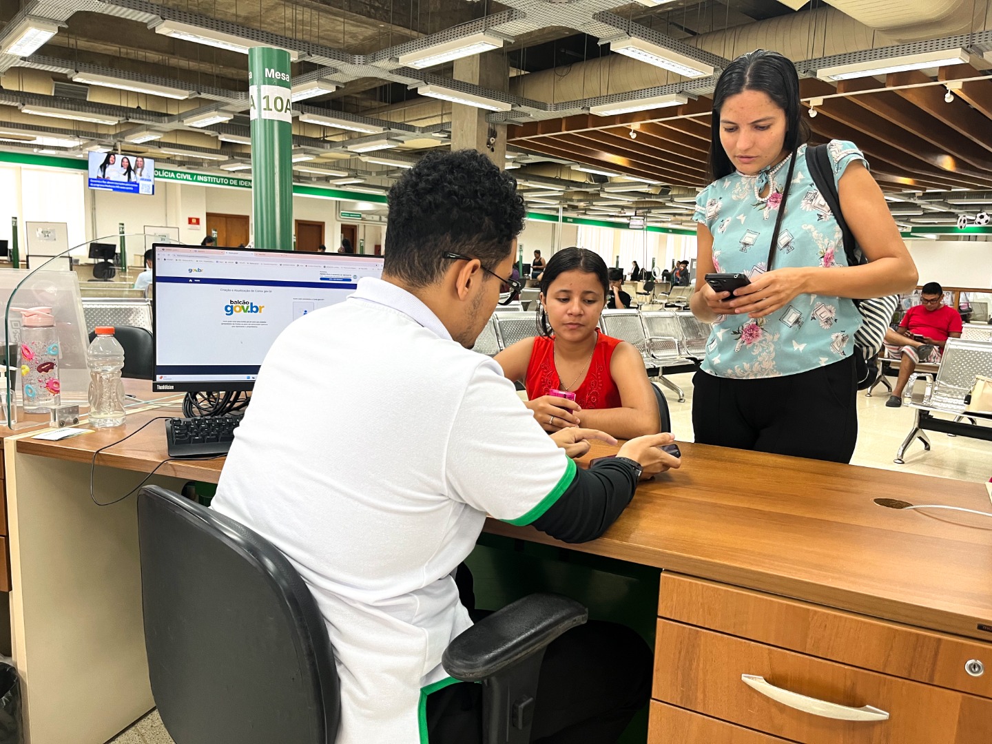 Atendentes da unidade OCA no Acre auxiliando cidadãos em guichês de atendimento, com pessoas aguardando e sendo orientadas em um espaço amplo.