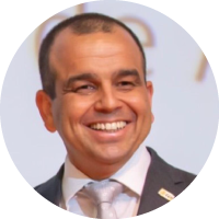 Foto de frente, da cintura para cima, do Valmir Gomes sorrindo. Ele possui cabelos castanhos, veste roupa paletó marrom claro e gravata prata.