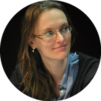 Foto de frente, da cintura para cima, da Miriamr, que possui cabelos claros, veste uma camisa branca de listras azuis, com um casaco preto por cima.