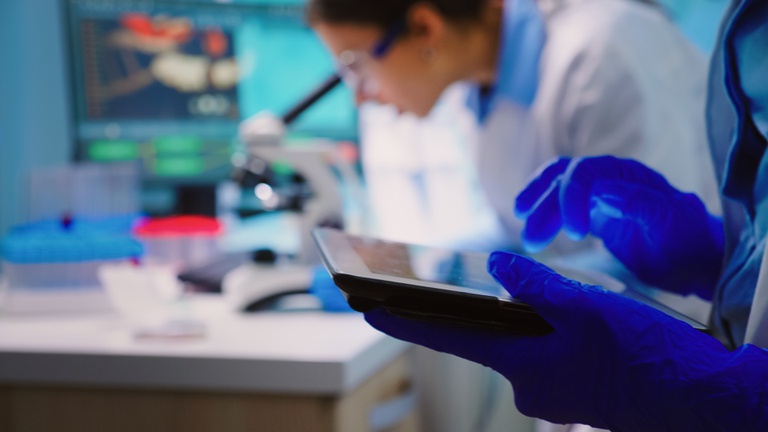 close-de-um-cientista-escrevendo-no-tablet-enquanto-uma-equipe-de-biologos-fazendo-pesquisas-biologicas-sob-o-microscopio-em-backgorund-tarde-da-noite.jpg