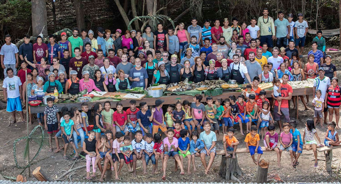 Foto de grupo grande, ao ar livre, sob árvores, reunindo dezenas de adultos e crianças de uma comunidade. Em primeiro plano, várias crianças sentadas em bancos de madeira. Atrás delas, um grupo maior de adultos em pé, alguns com aventais, ao redor de uma longa mesa coberta de folhas de bananeira com comida servida, parece uma refeição comunitária ou celebração coletiva. Todos posam sorrindo para a foto.