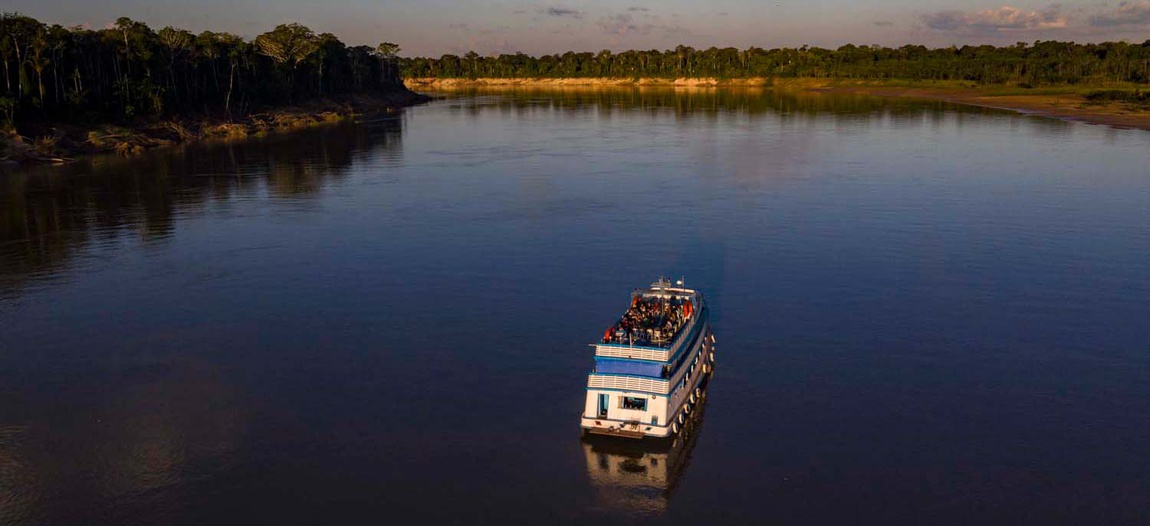 Fotografia aérea de um grande barco branco com detalhes azuis, com dois andares, navegando sozinho no centro de um rio amazônico largo e calmo; há pessoas reunidas no convés superior. As margens do rio são cobertas por floresta densa de um lado e por uma faixa de praia arenosa do outro. É um fim de tarde com poucas nuvens no céu.