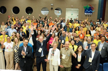Grande grupo de participantes posa para foto em um auditório, com várias pessoas em pé, usando crachás e algumas com uniformes amarelos ou laranja. Parte do grupo levanta os braços ou acena para a câmera. Ao fundo, aparecem cadeiras azuis, portas de acesso e a marca do Ibama com a sigla MMA na parede lateral.