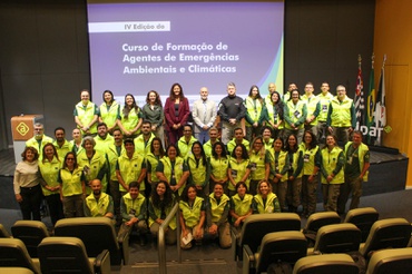Um grupo de 48 pessoas posa para foto coletiva em frente a uma tela de projeção em um auditório. A maioria usa colete amarelo fluorescente sobre roupas verdes ou escuras, com crachás pendurados no pescoço. Algumas pessoas estão em pé em fileiras e outras estão ajoelhadas ou sentadas à frente. No fundo, a tela exibe o texto: “IV Edição do Curso de Formação de Agentes de Emergências Ambientais e Climáticas”. À direita da imagem, há bandeiras em suportes e parte da marca da Ambipar. À frente do grupo, aparecem fileiras de poltronas do auditório. O ambiente é amplo, com iluminação artificial e paredes em tons escuros e amarelos.
