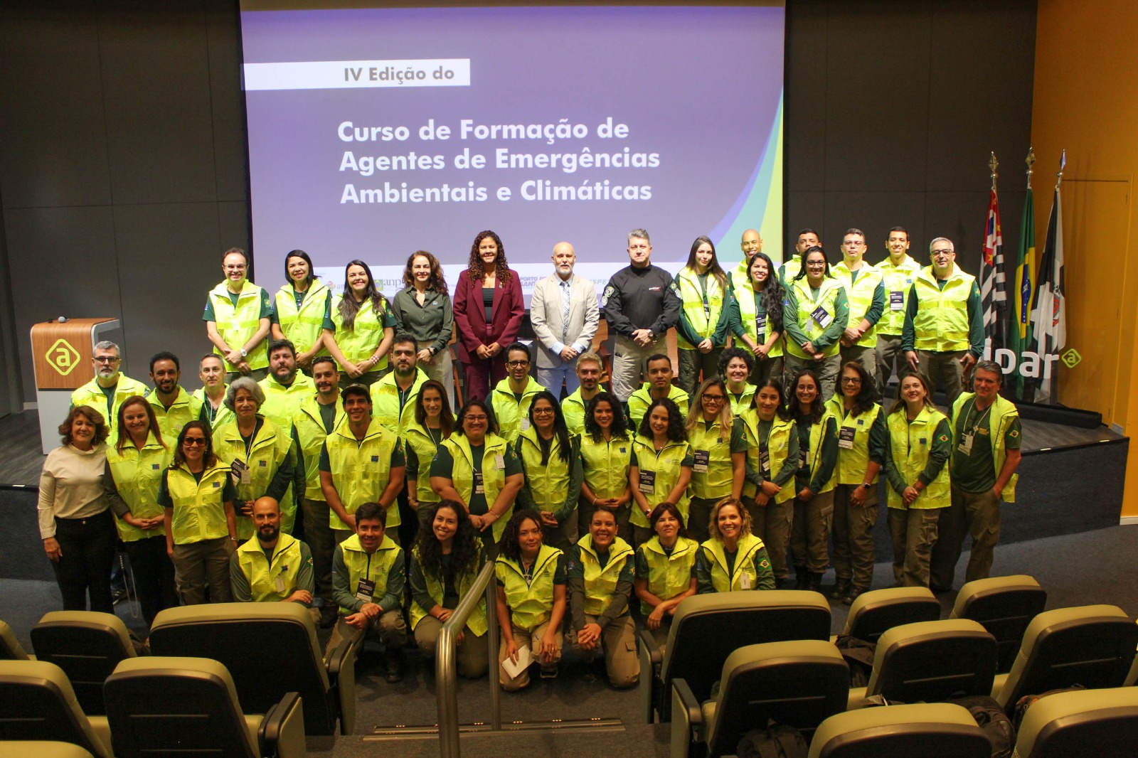 Um grupo de 48 pessoas posa para foto coletiva em frente a uma tela de projeção em um auditório. A maioria usa colete amarelo fluorescente sobre roupas verdes ou escuras, com crachás pendurados no pescoço. Algumas pessoas estão em pé em fileiras e outras estão ajoelhadas ou sentadas à frente. No fundo, a tela exibe o texto: “IV Edição do Curso de Formação de Agentes de Emergências Ambientais e Climáticas”. À direita da imagem, há bandeiras em suportes e parte da marca da Ambipar. À frente do grupo, aparecem fileiras de poltronas do auditório. O ambiente é amplo, com iluminação artificial e paredes em tons escuros e amarelos.